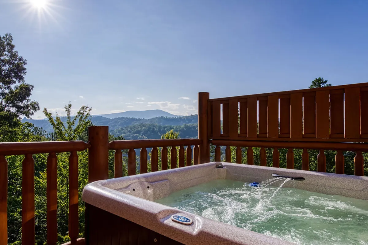 An exterior deck with a bubbling hot tub overlooking the majestic Smoky Mountains. Wooden railings frame the view, inviting guests to relax and soak in the scenery, day or night.