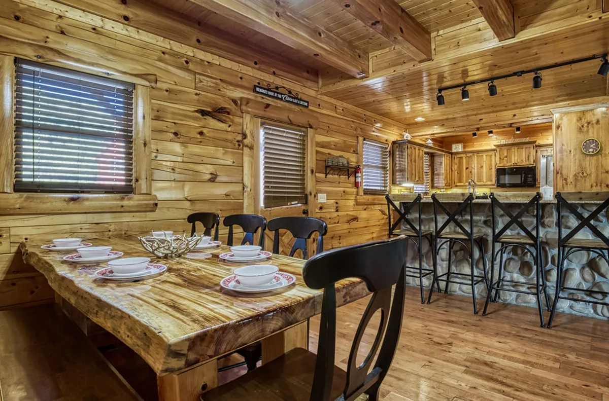 A cozy dining area with a rustic wooden table, stone bar counter, kitchen cabinetry, and large sliding windows with wooden blinds. The space invites gatherings, meals, and lively conversations.