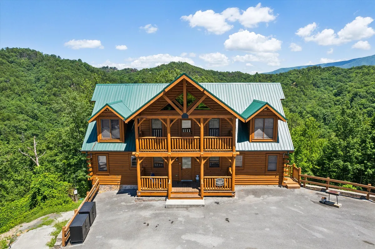 A stunning log cabin with a green metal roof is nestled atop a ridge, surrounded by the rolling peaks of the Smoky Mountains. The spacious porch offers sweeping sunset views, inviting guests to unwind and take in the natural beauty. The cabin's warm wood exterior and elevated position exude both luxury and rustic charm.