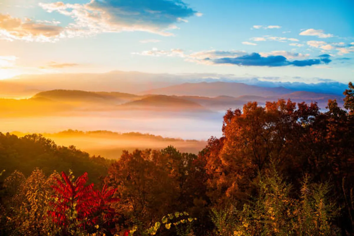 A wide view of a breathtaking mountain range under a sky tinged with soft colors, with rolling hills and colorful foliage filling the foreground. The misty valley adds a sense of depth and tranquility, perfectly representing the awe-inspiring vistas the Smoky Mountains are known for.
