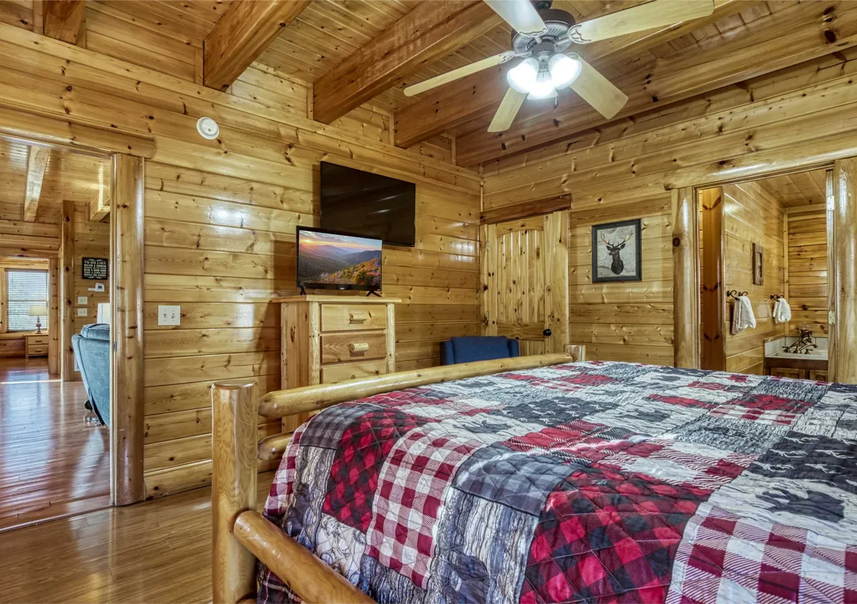 A cozy bedroom featuring a rustic log bed with a patchwork quilt, wooden walls, a TV, and an armchair by the window. Natural light filters in, creating a warm and inviting atmosphere ideal for relaxation or reading.