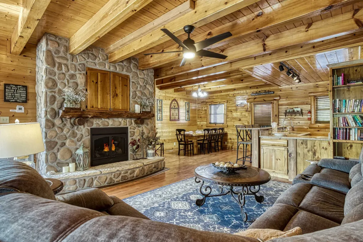 This inviting family room features a central stone fireplace, exposed wooden beams, a plush sofa, a dining area, and a large blue area rug. The warm wood finishes and open layout create a welcoming space for gatherings, with the fireplace serving as a natural focal point for cozy evenings.