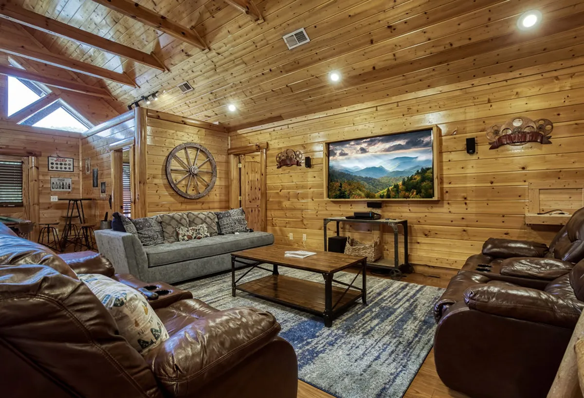 A cozy living area with comfortable sofas arranged around a coffee table, a large TV mounted on wood-paneled walls, and a decorative wagon wheel. The warm, golden lighting and ample seating highlight the room's suitability for movie nights and relaxed socializing.