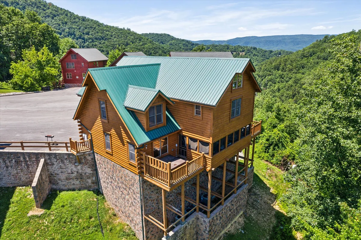 This exterior shot shows a charming log cabin with a green metal roof, a large deck area, and sweeping views of the surrounding mountains. The scene is set for outdoor gatherings with deck seating, ideal for grilling and dining while enjoying the stunning natural scenery.