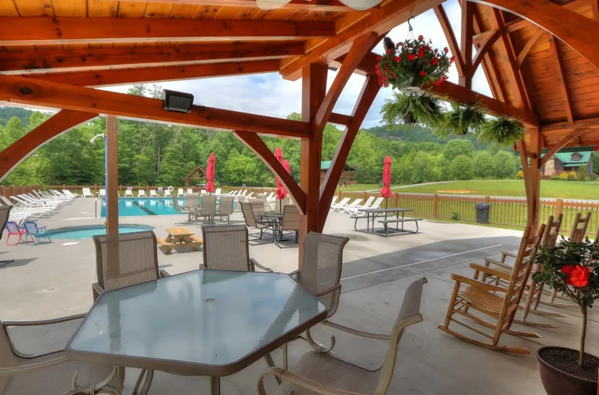 A lively scene featuring a community swimming pool surrounded by red umbrellas, a wooden gazebo, and rocking chairs, with friends and families enjoying the water and socializing. The setting exudes a cheerful, communal atmosphere ideal for memorable group experiences.