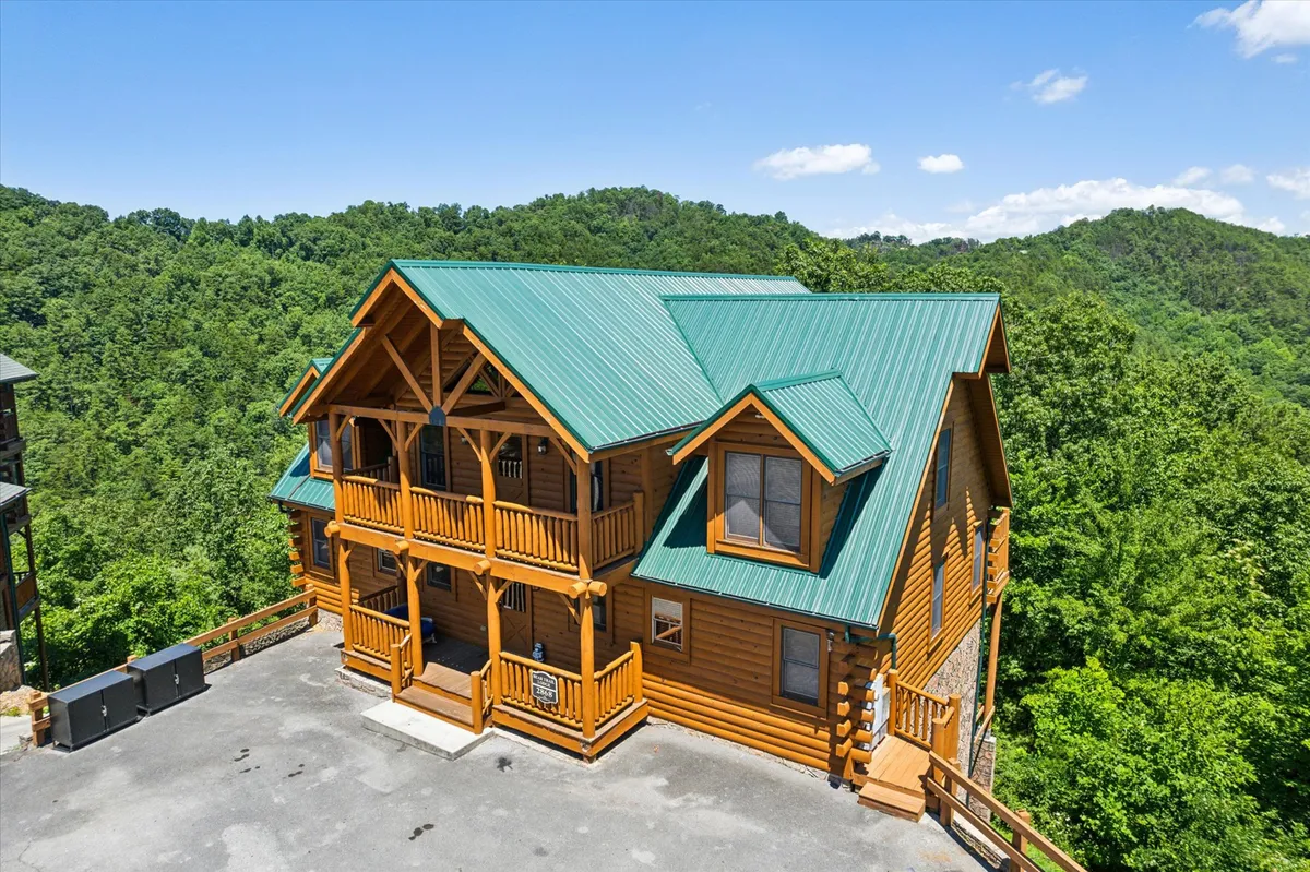 A picturesque log cabin with a green metal roof is nestled among trees, highlighted by inviting balcony railings and surrounded by the tranquility of the mountains. The cabin’s exterior exudes exclusivity and is ideal for romantic getaways within a prestigious mountain community.